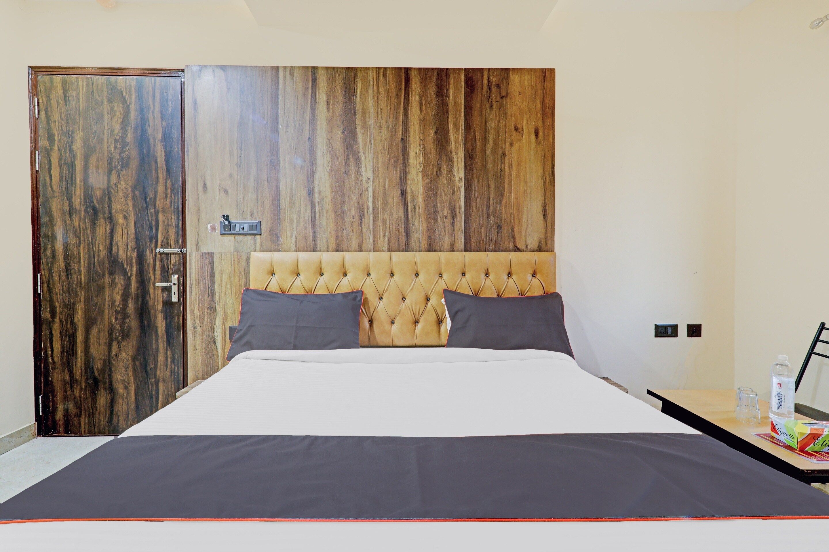 Un dormitorio con cabecera de madera, una cama con edredón blanco y oscuro, dos almohadas, una mesita de noche con una botella de agua y una caja de pañuelos, y una puerta con cerradura.