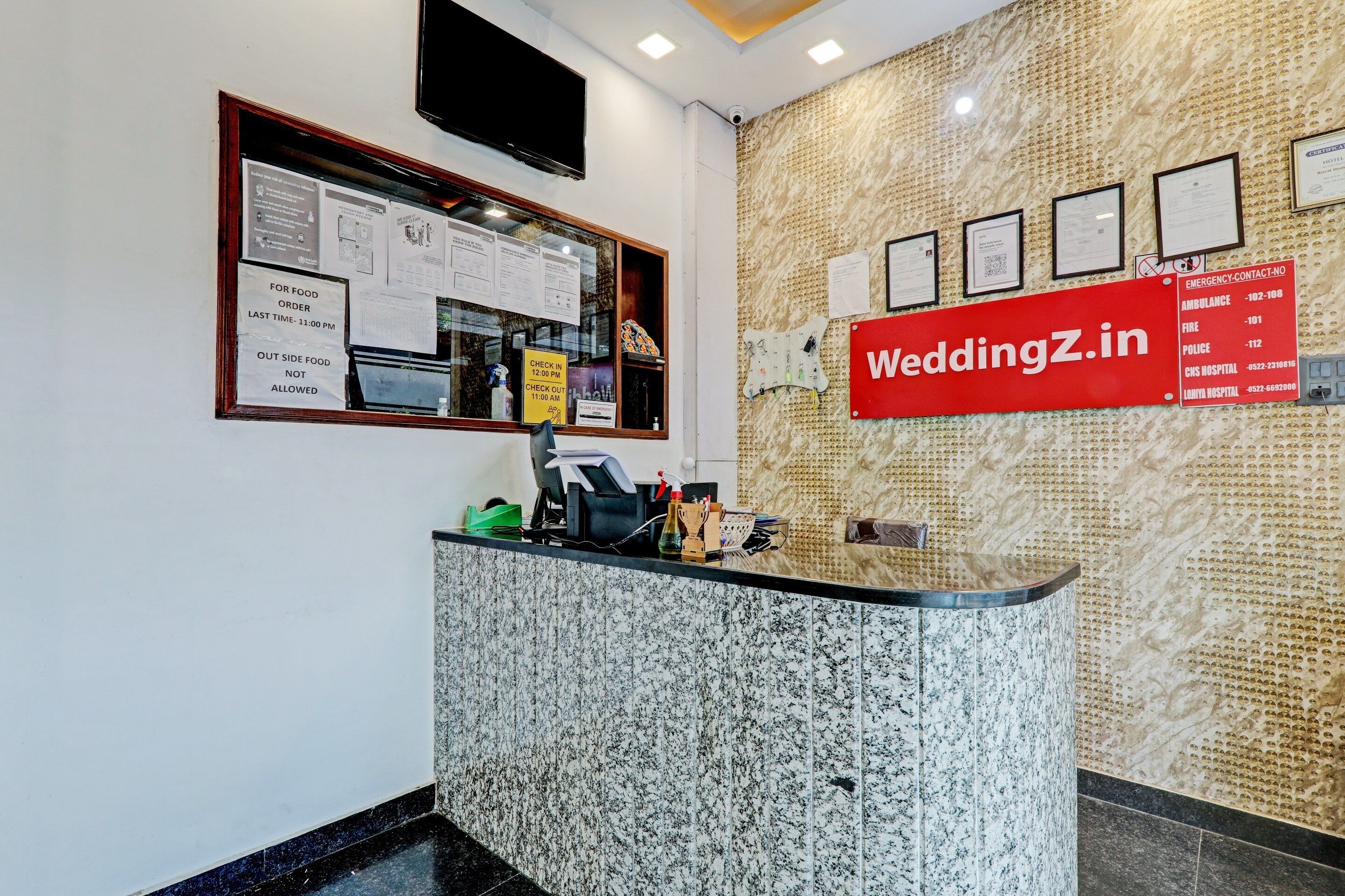 A hotel reception desk with a marble counter, a mounted television, and various informational posters.