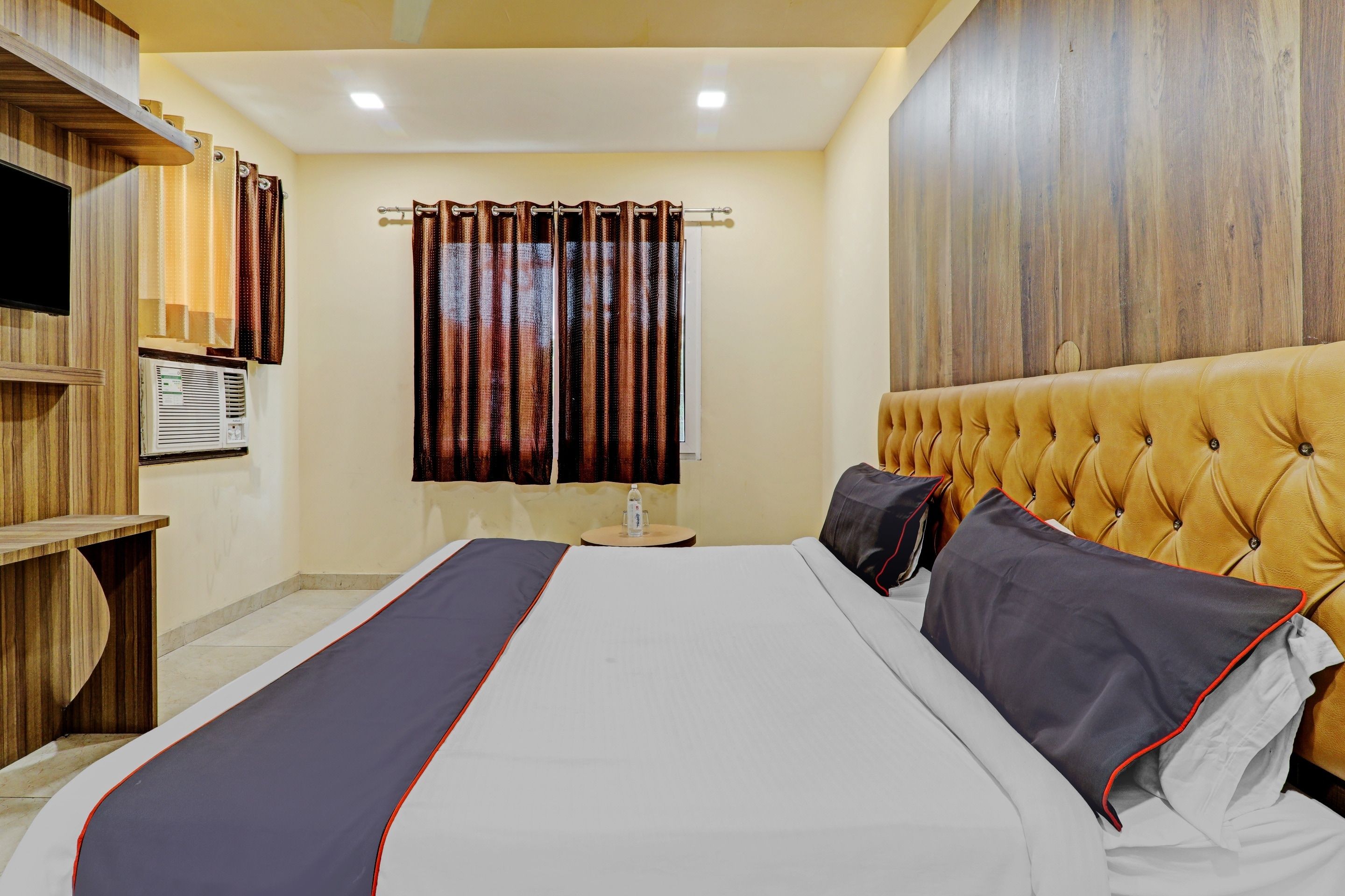 A hotel room with a bed, a wooden headboard, a window with curtains, a small table with a bottle, and a wall-mounted TV.