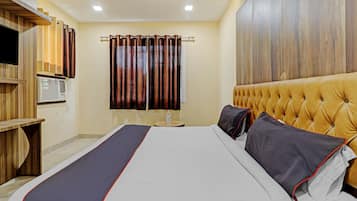 A hotel room with a bed, a wooden headboard, a window with curtains, a small table with a bottle, and a wall-mounted TV.