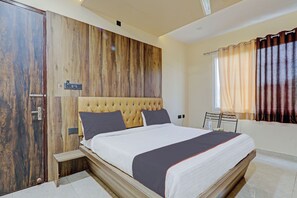 A bedroom with a wooden panel wall, a tufted headboard, a bed with white and dark bedding, a bedside table, and a window with curtains. - Townhouse Jayati Oasis Inn (Lucknow)