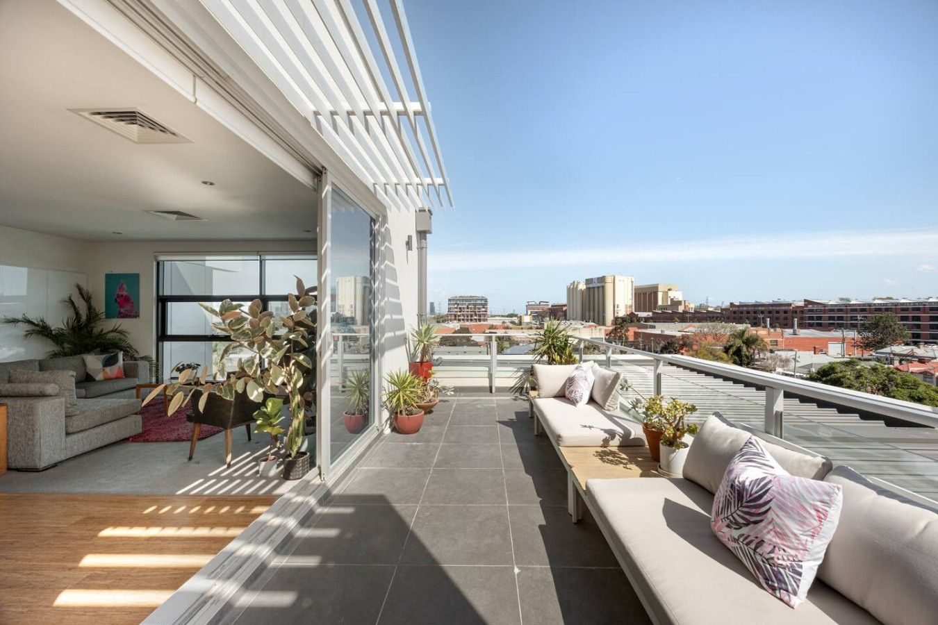 Una sala moderna con sofá y comedor, conectada a un balcón con plantas en macetas y vistas a la ciudad.
