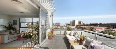 Una sala moderna con sofá y comedor, conectada a un balcón con plantas en macetas y vistas a la ciudad.