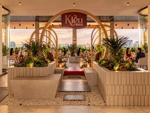 Un espacio interior moderno con un arco central identificado como 'Kiêu by KITA', rodeado de zonas de estar con plantas y vistas a la ciudad.
