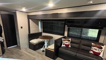 Un espace de vie compact comprenant un coin cuisine, un canapé agrémenté de coussins décoratifs et une fenêtre avec des stores.