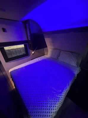 A bed with a patterned bedspread, a wall-mounted heater, and a small shelf with a lamp. - Cozy RV  close to bird sanctuary with chill vibes and fantastic sunset views. (San Ygnacio)