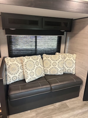 A dark brown leather sofa with patterned cushions, a window with blinds, and a wooden wall. - Cozy RV  close to bird sanctuary with chill vibes and fantastic sunset views. (San Ygnacio)
