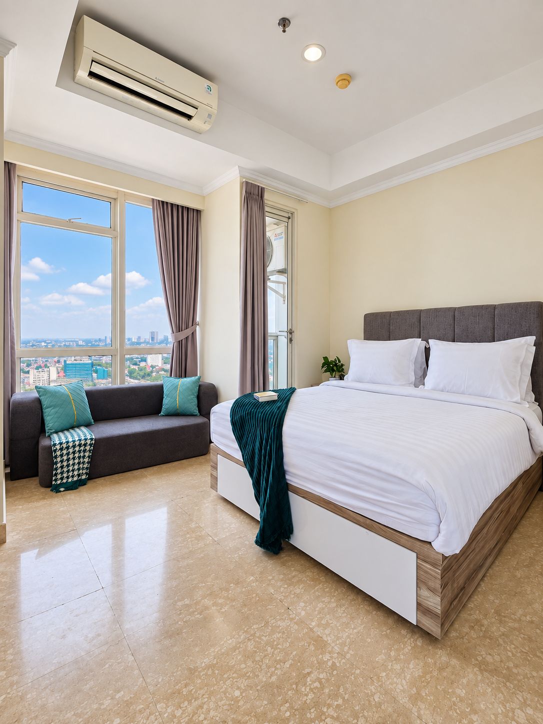 A hotel room with a bed, a sofa, a window with curtains, an air conditioning unit, and a view of the city.