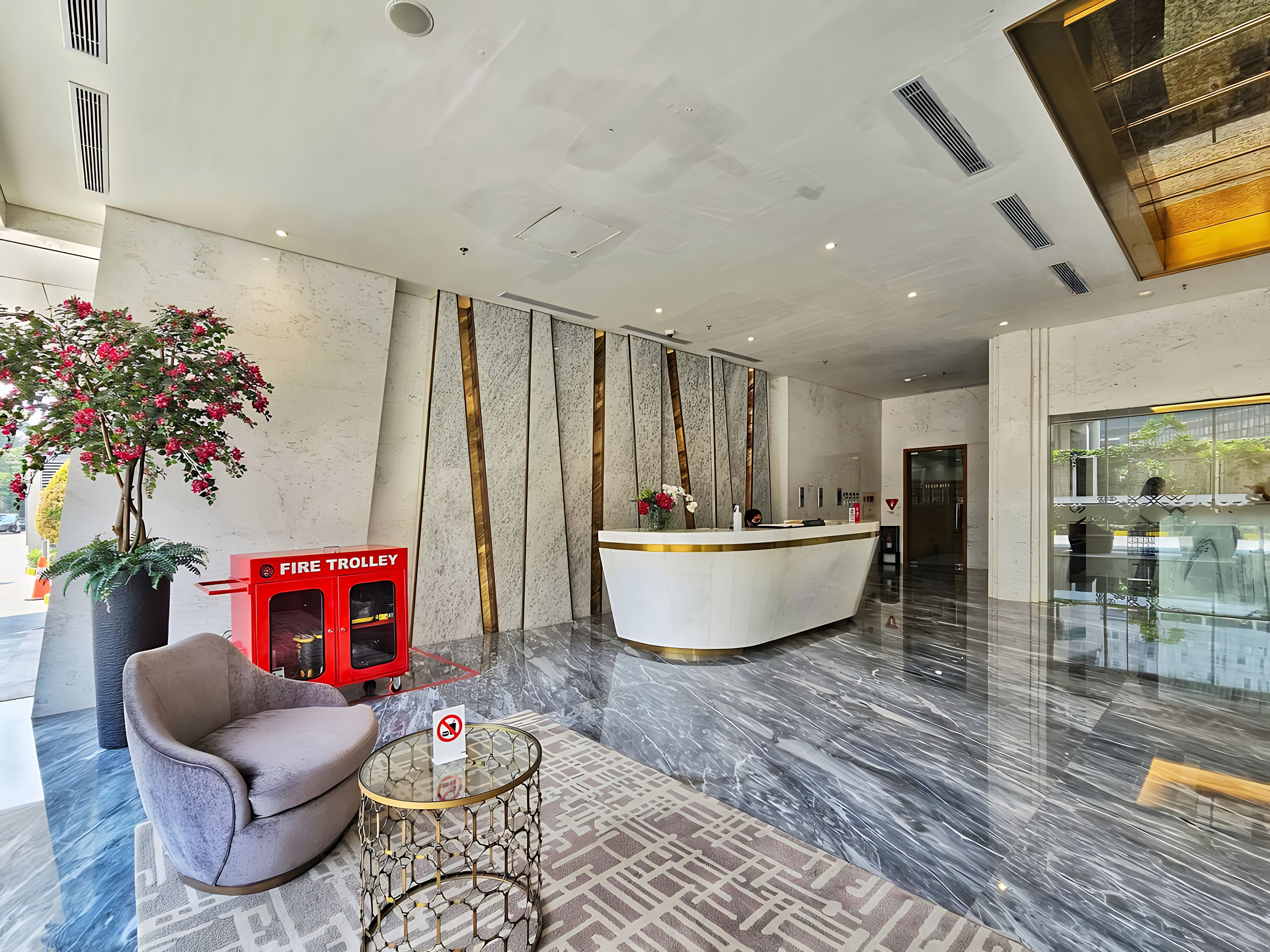 Una zona de recepción moderna con suelo de mármol, mostrador de recepción, un carrito rojo, una planta en maceta y una sala de estar con una mesa de vidrio.