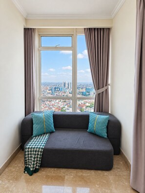 A corner room with a city view, a dark grey sofa with teal pillows, and a patterned throw. - iNDIGO - Studio - Menteng Park (Jakarta)