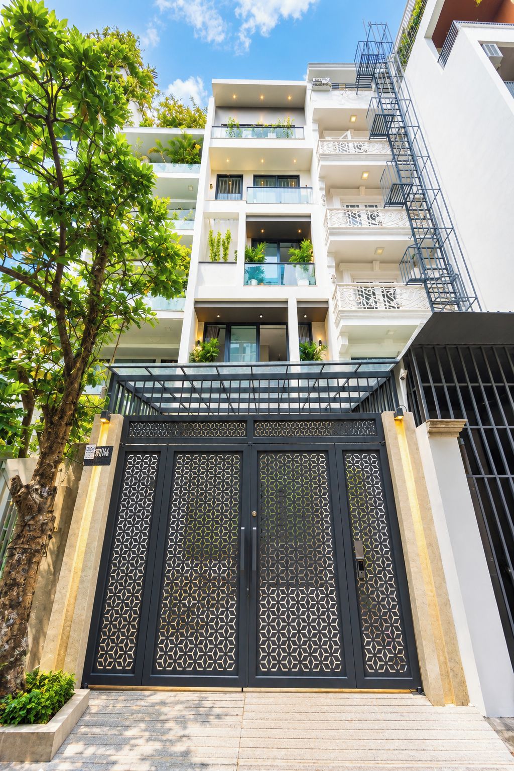 A multi-story residential building with a gated entrance, balconies with plants, and a staircase leading to the entrance.