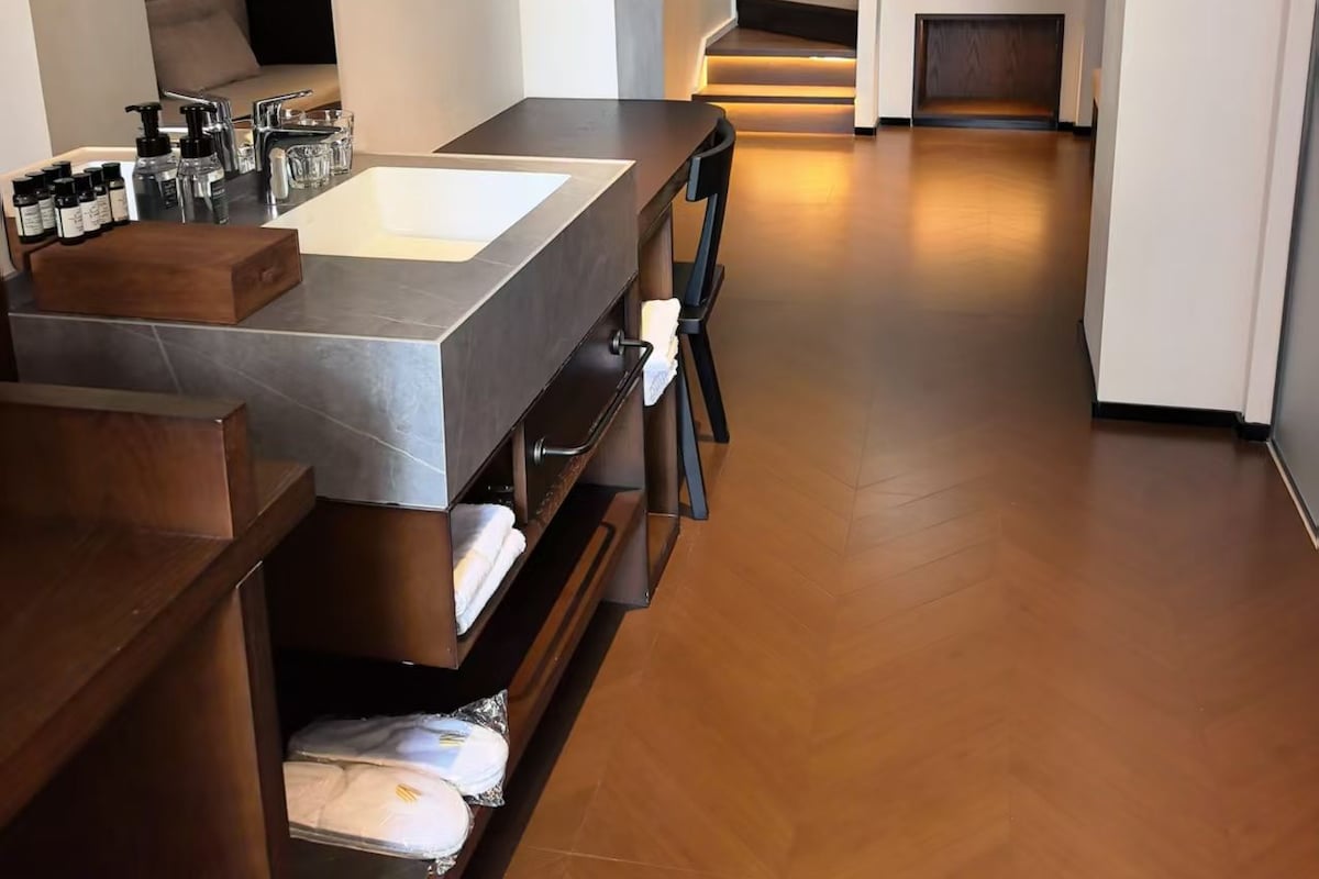 A modern bathroom with a large sink, mirror, and wooden vanity. There is a staircase leading to a bedroom with a bed and a window.