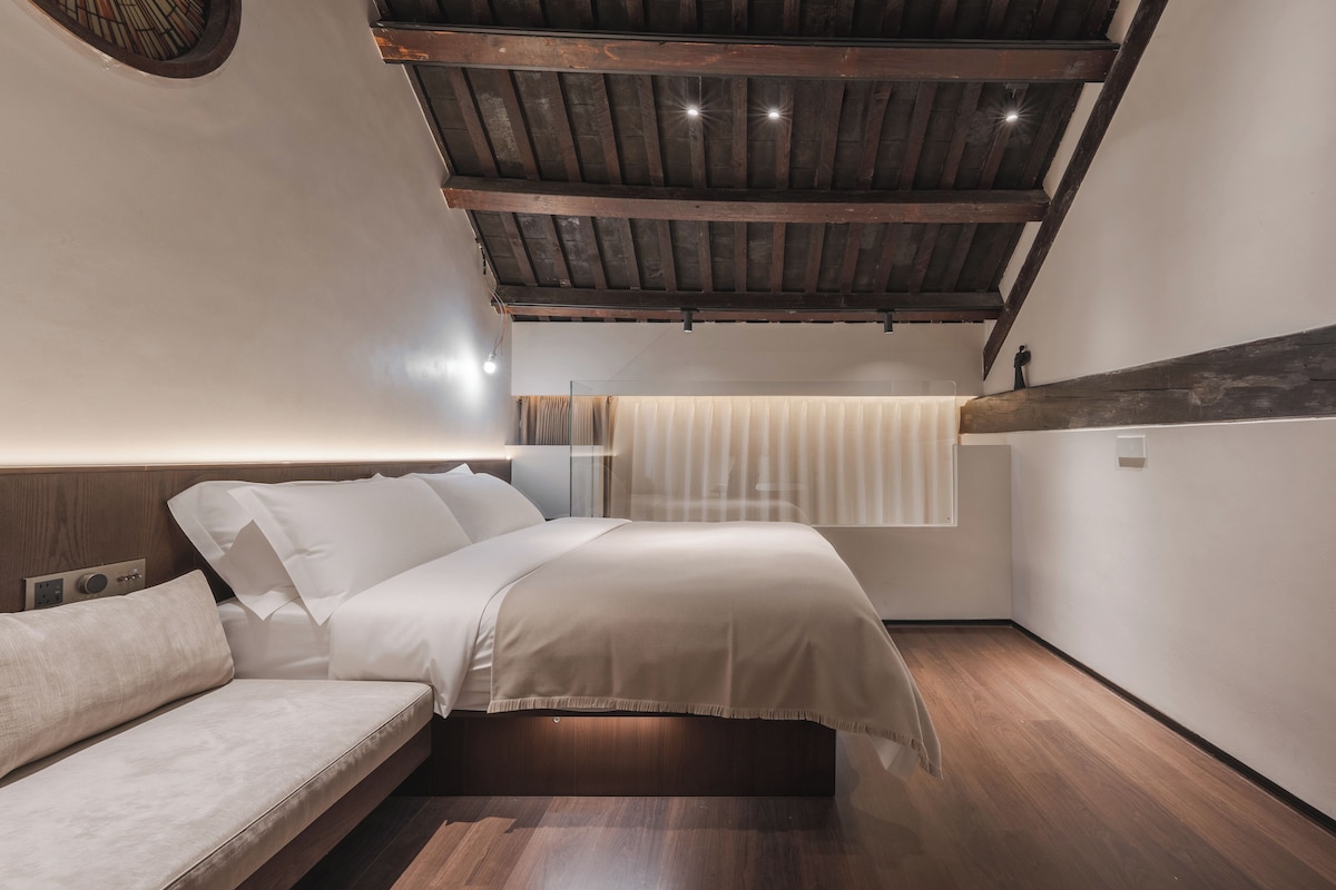 A bedroom with a wooden ceiling, a bed with white bedding, a wooden floor, and a window with curtains.