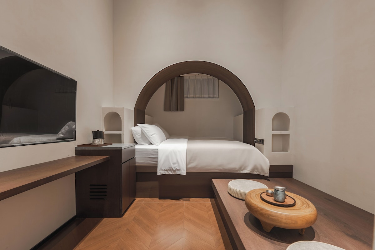 A compact hotel room with a bed, a TV, a small table, and a wooden bench.