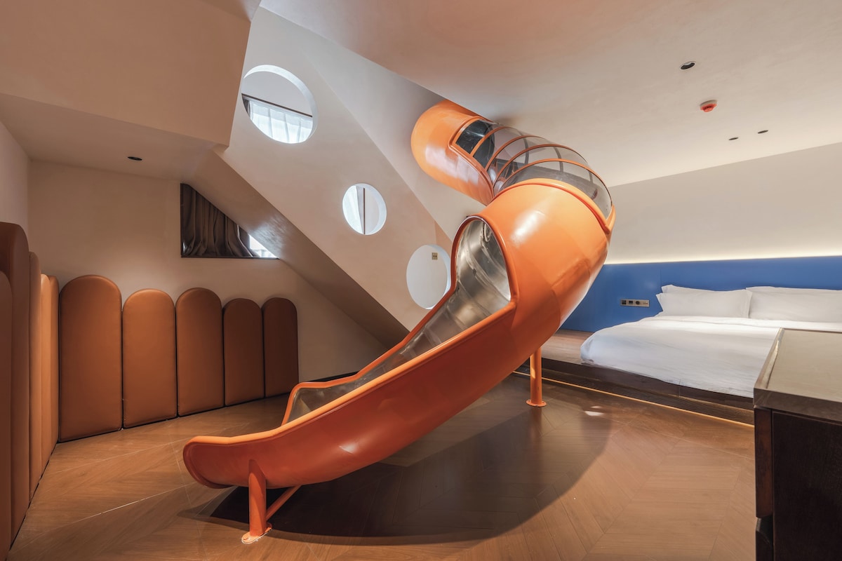 A modern hotel room with a unique orange slide, a bed, and a large window.