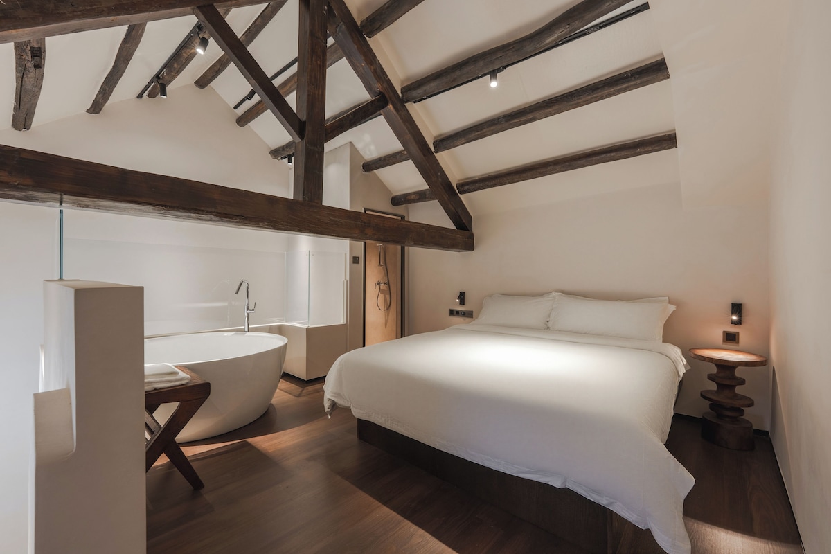 A bedroom with a large bed, a bathtub, wooden beams on the ceiling, and a small bedside table with a lamp.