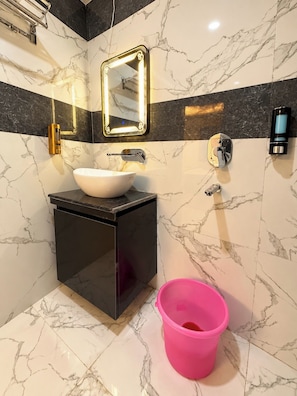 A bathroom with a white sink, black cabinet, marble walls, a mirror with lights, and a pink bucket.