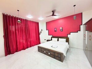 A bedroom with a bed, a ceiling fan, a red wall, and a window with curtains. - Adiva Villa By Vintara Stays (Murud)