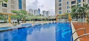 Una piscina con cubierta de madera, rodeada de vegetación y edificios modernos al fondo.