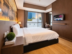 A modern hotel room with a large bed, a flat-screen TV, a desk, and a window with a city view. - Kisah By SanLiving - 3BR - Kasablanka (Jakarta)