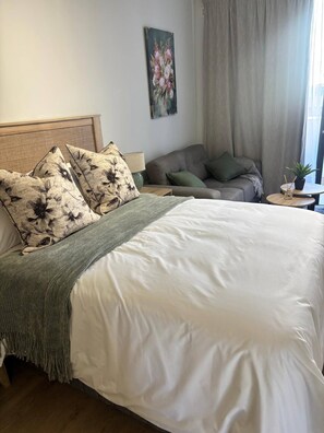 A neatly made bed with a white duvet and floral pillows, a grey sofa with cushions, a small round table with a plant, and a painting on the wall. - Rosebank Retreat, Business or Leisure (Johannesburg)