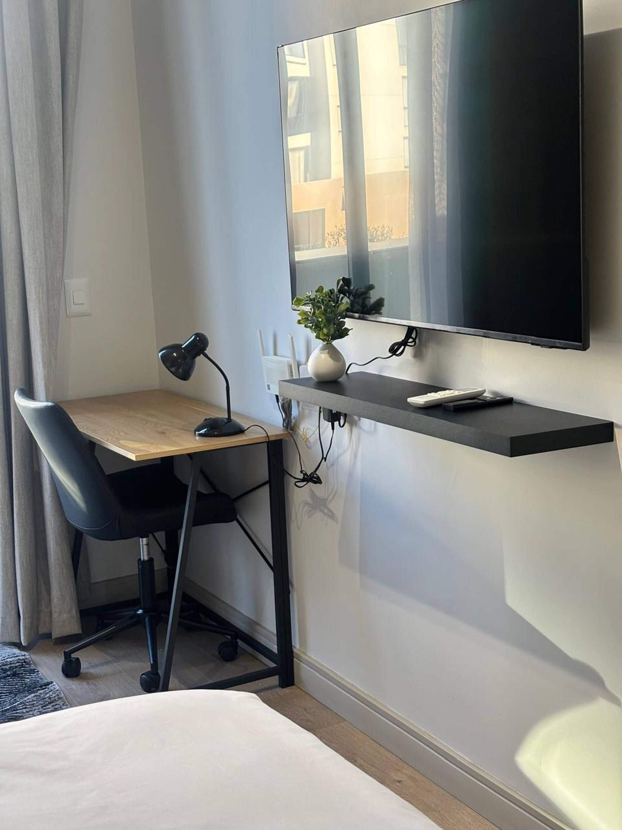 A modern hotel room with a desk, chair, flat-screen TV, and a bedside table.