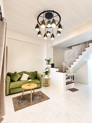 A modern living room with a green sofa, a round wooden coffee table, and a patterned rug. A staircase with a metal railing leads to an upper level. - Charming 2-bedroom House Rental in Muang Pattaya (Pattaya)