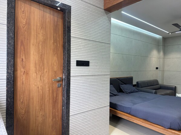 A modern room with a wooden door, a bed with a dark blue bedspread, a grey sofa, and a ceiling fan.