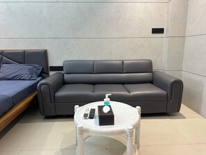 A modern room with a grey sofa, a small white table with a remote control, and a bed with a grey mattress.