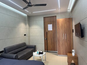 A modern hotel room with a sofa, a small table with a remote control, a wooden wardrobe, and a ceiling fan.