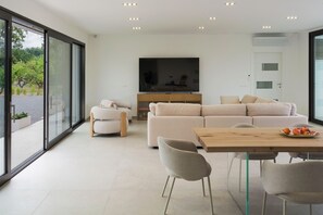A modern living room with a large flat-screen TV, a sofa set, a dining table with chairs, and a large window offering a view of greenery. - Stunning villa with private pool in Taormina - DiCasaInSicilia (Taormina)