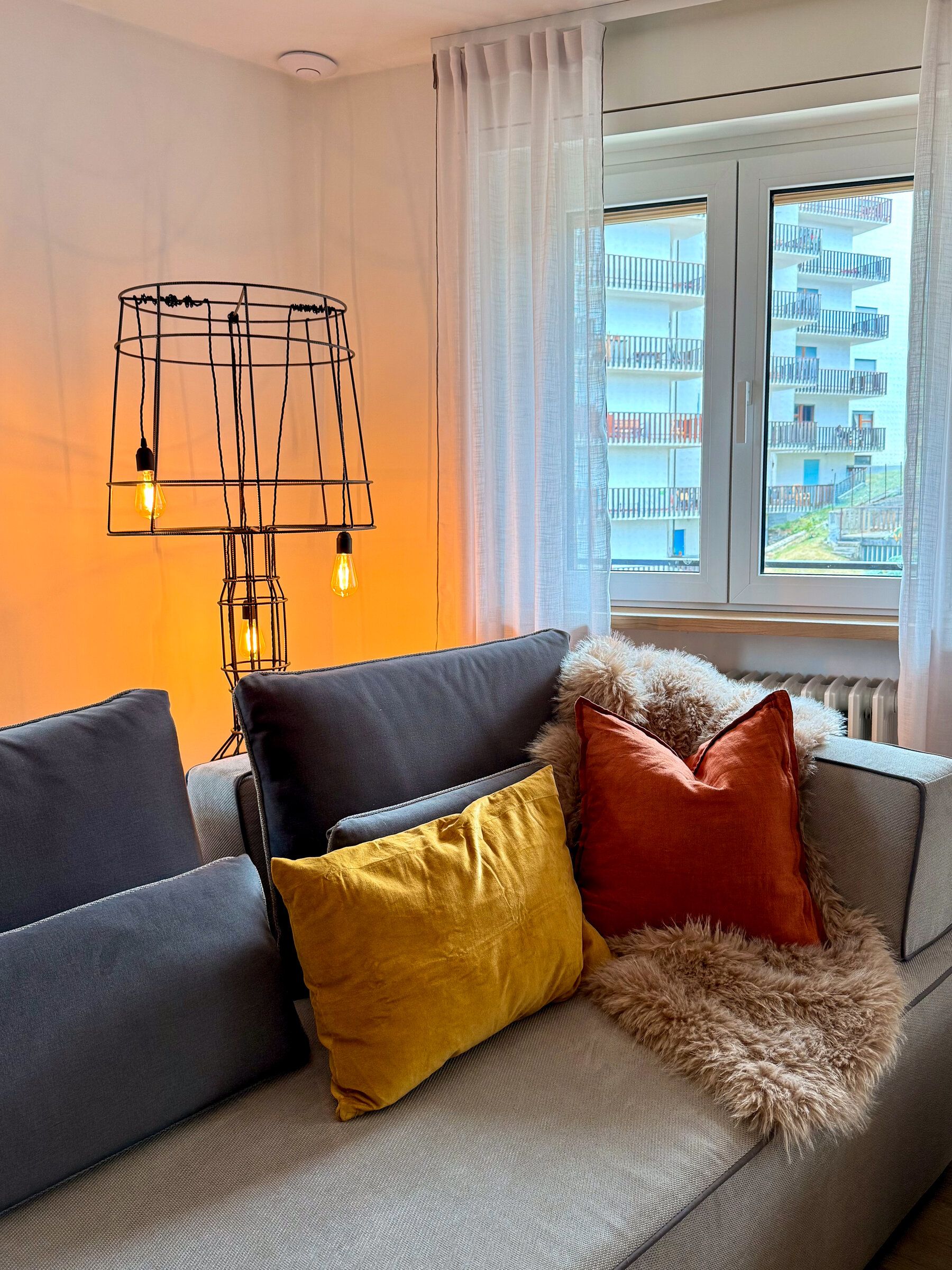 A modern living room with a sofa, decorative pillows, a unique light fixture, and a view of a building through a window.