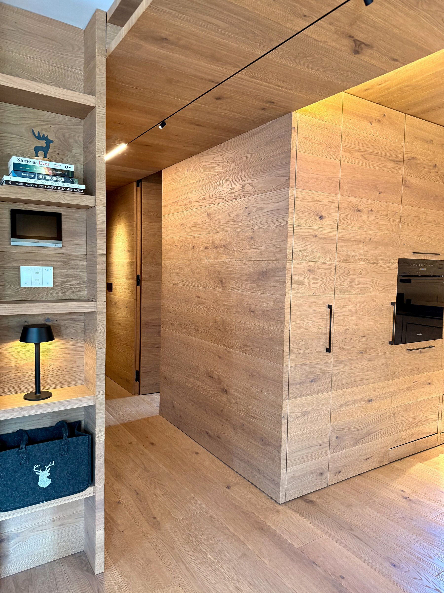 A modern kitchen with wooden cabinetry, a built-in microwave, and a shelf with books.
