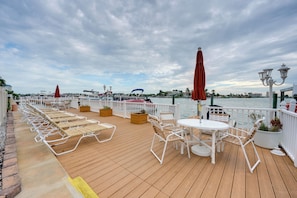 A deck with sun loungers, tables, and umbrellas, overlooking a body of water with buildings in the background. - Bayside Bliss | First-Floor Condo with Water Views (St. Pete Beach)