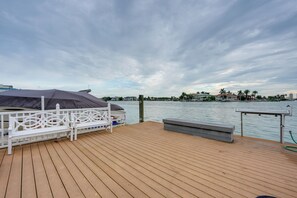 A wooden deck with a white railing, a covered seating area, and a view of a calm body of water with buildings in the background. - Bayside Bliss | First-Floor Condo with Water Views (St. Pete Beach)