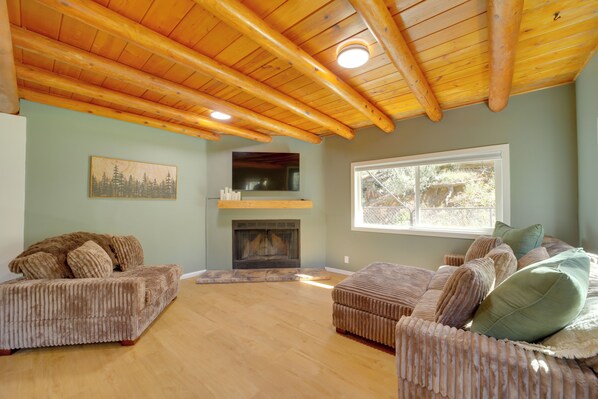 A spacious living room with a high wooden ceiling, a fireplace, and a large window. - Cozy Prescott Cabin, Surrounded By Ponderosa Pines (Prescott)