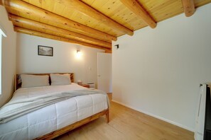 A bedroom with a wooden bed frame, a white mattress, and a blanket. There is a bedside table with a lamp and a wall-mounted light. The room has wooden flooring and a wooden ceiling with exposed beams. - Cozy Prescott Cabin, Surrounded By Ponderosa Pines (Prescott)