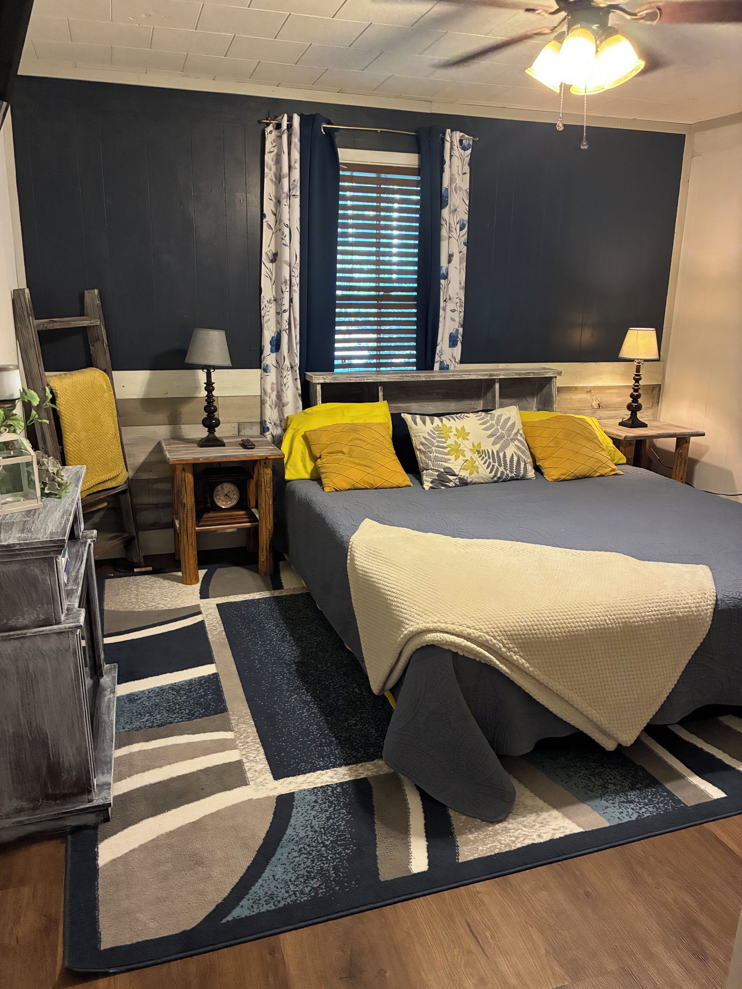 A bedroom with a bed, nightstands, a lamp, a window with curtains, a ceiling fan, and a wooden ladder.