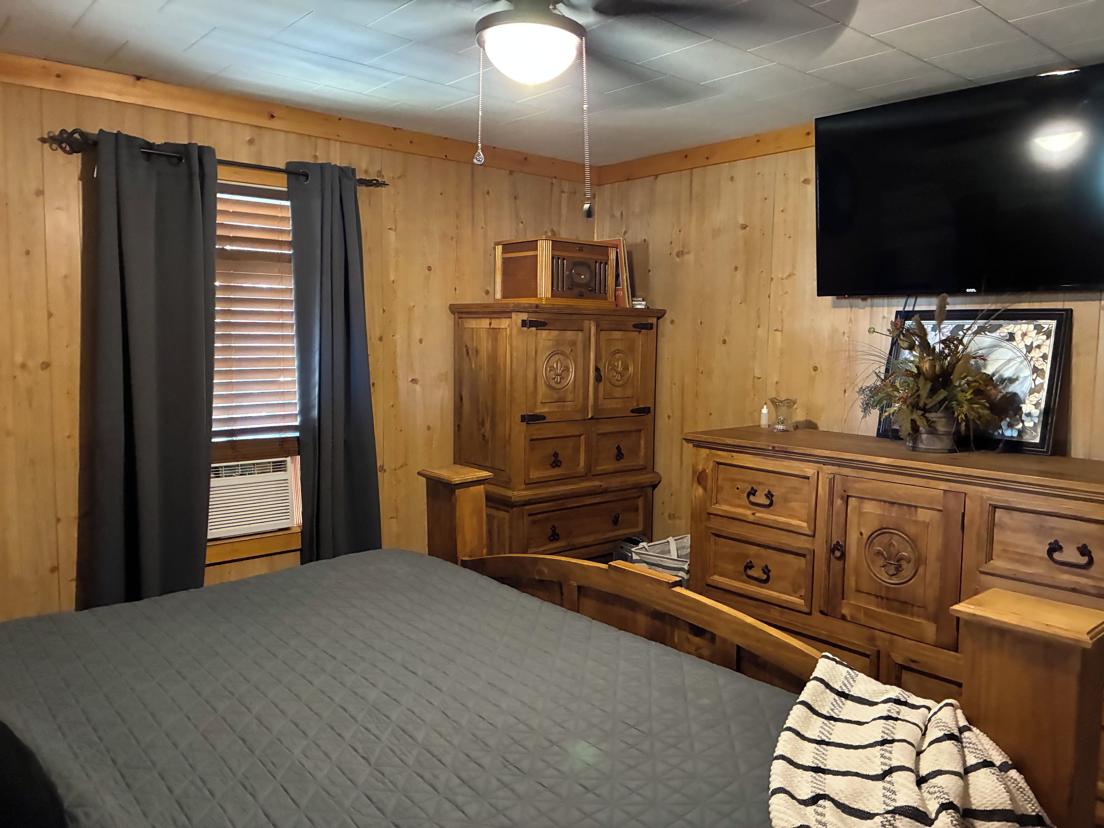 Une chambre avec un lit, une commode en bois, une télévision et une fenêtre avec des rideaux.