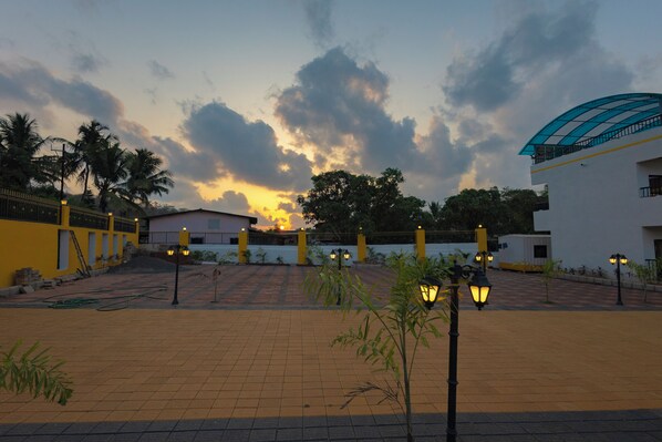 Un patio con un camino pavimentado, un edificio con techo curvo y una puesta de sol al fondo.