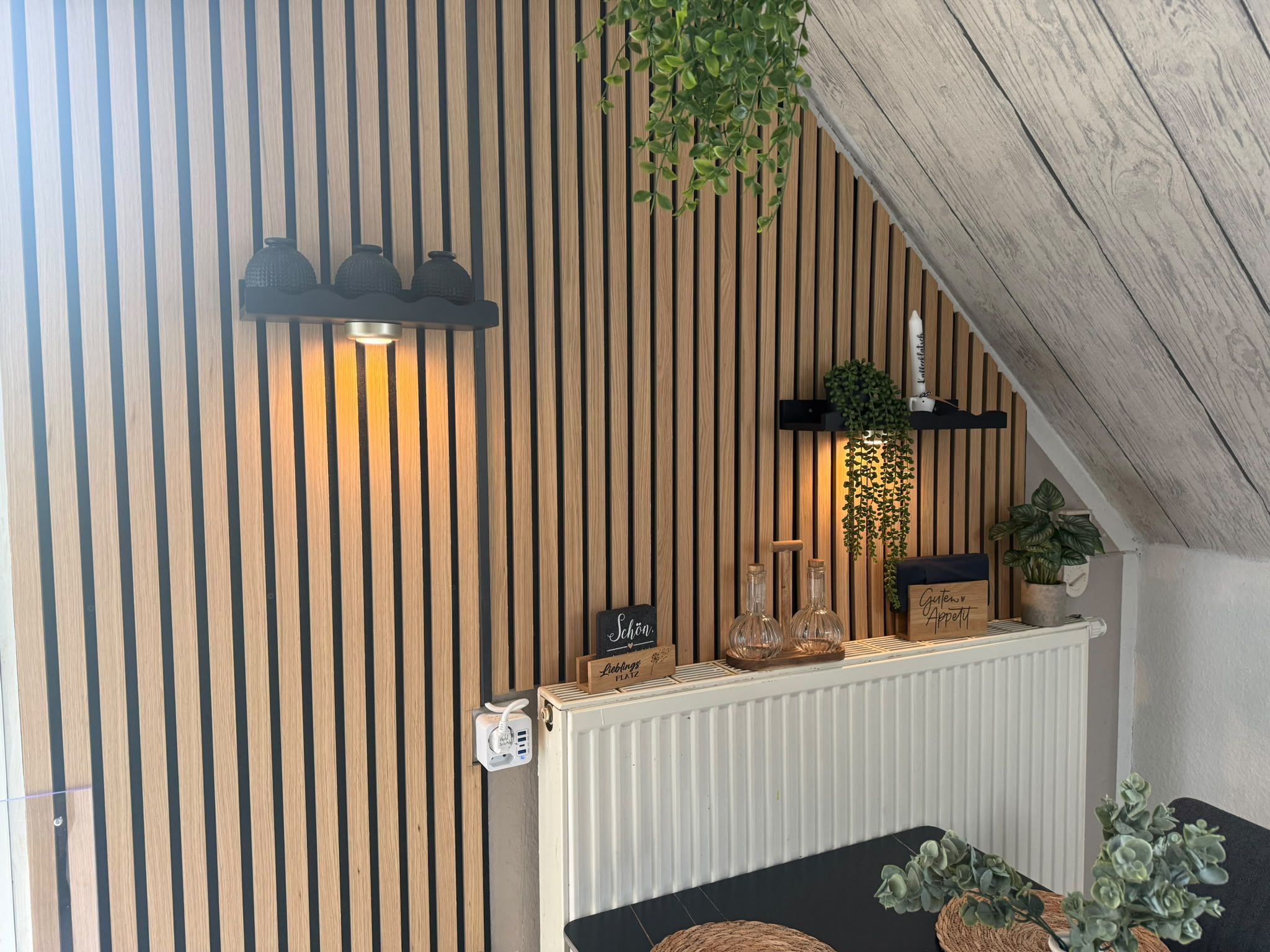 Ein Zimmer mit einer horizontalen Holzwand, einem weißen Heizkörper, einem schwarzen Tisch mit einer gewebten Tischunterlage und einer hängenden Pflanze.