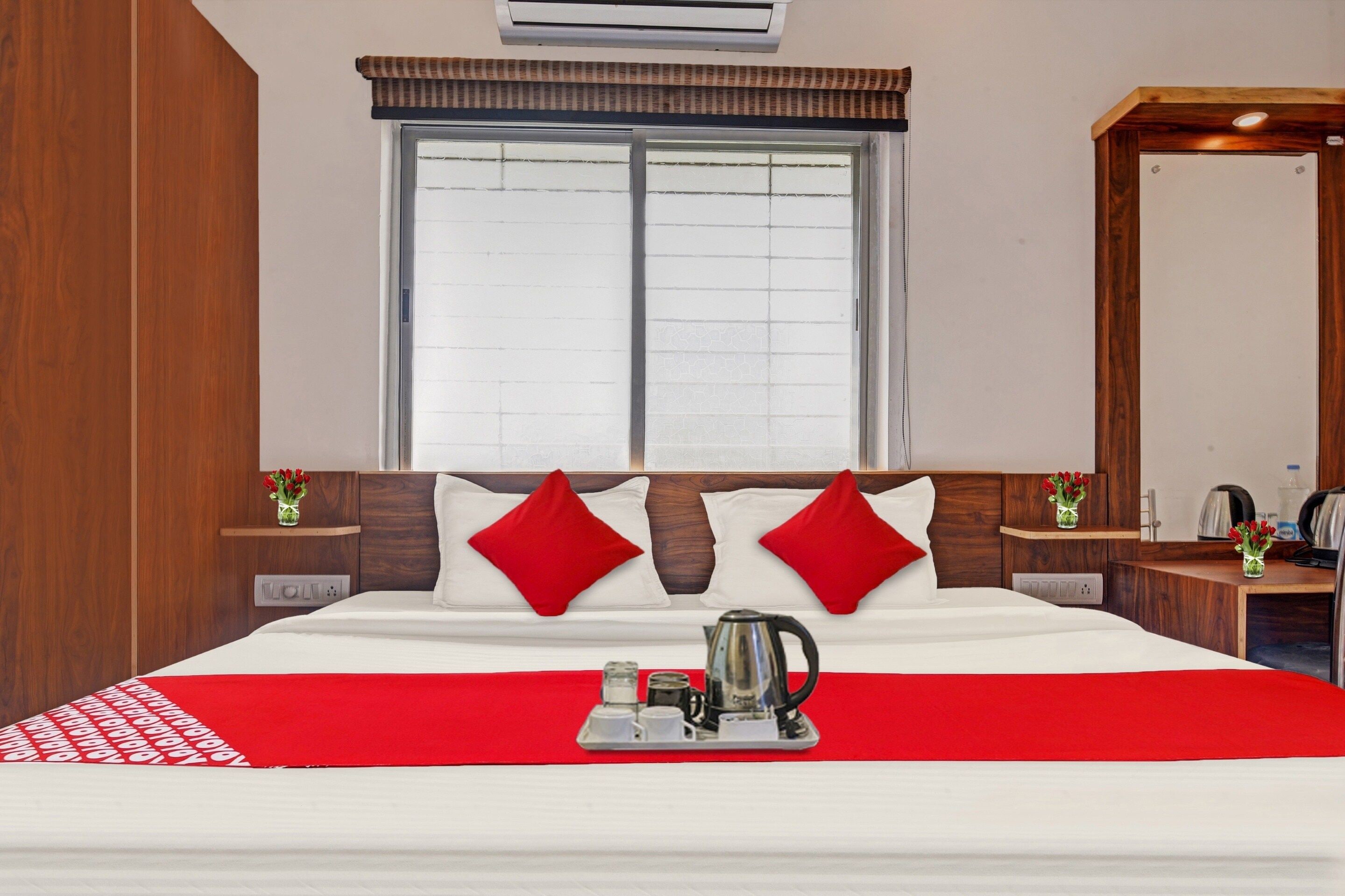 Una habitación de hotel con una cama grande, una ventana con persianas, una mesita de noche con un florero y un espejo en la pared.