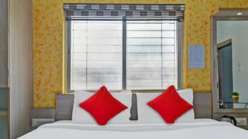 A hotel room with a large bed, two red pillows, a window with blinds, and a mirror on the wall.