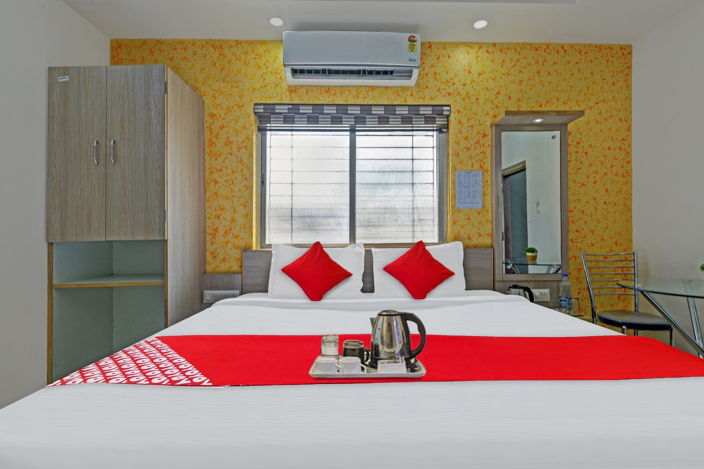 A hotel room with a bed, a wardrobe, a window with blinds, a mirror, and a table with a kettle.