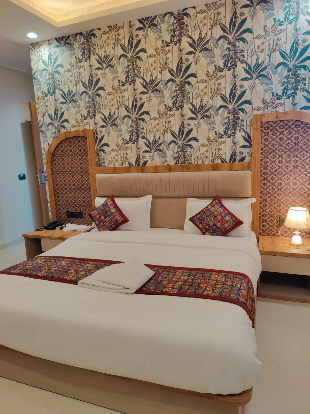 Una camera d'albergo con un letto grande, una testiera in legno, carta da parati decorata e un comodino con una lampada.