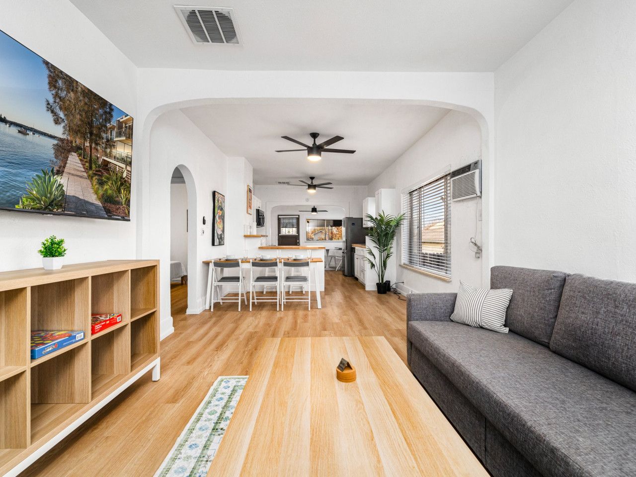 A modern living room with a grey sofa, wooden flooring, a dining area with a table and chairs, and a view of a body of water through an arched window.