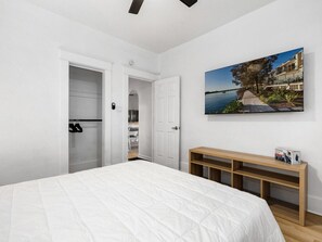 A bedroom with a large bed, a wooden bench, a door leading to a bathroom, and a painting of a coastal scene. - Casitas en Pueblo - Washington House (Pueblo)