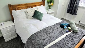 Ein ordentlich bezogenes Bett mit einer grauen, strukturierten Decke, weißen Kissen und einem grünen Akzentkissen. Ein weißer Nachttisch mit einer Lampe, einer kleinen Topfpflanze und einer Wasserflasche auf dem Boden neben dem Bett.