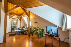 Une pièce avec un plafond en pente, des poutres en bois, un téléviseur posé sur un meuble, une plante et une vue sur un bâtiment par la fenêtre.