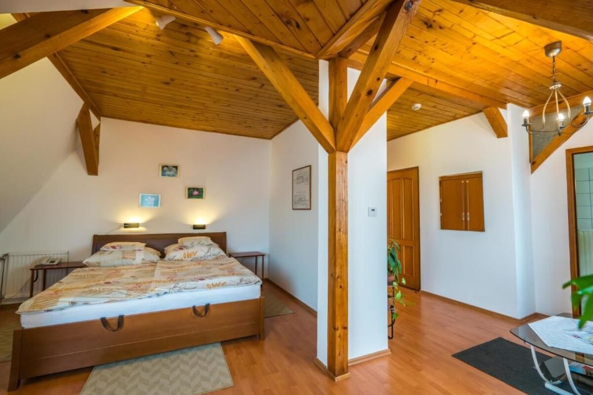 Une chambre avec un plafond en bois, un lit recouvert d’une couvre-lit à motifs, une table de chevet et une lampe fixée au mur.